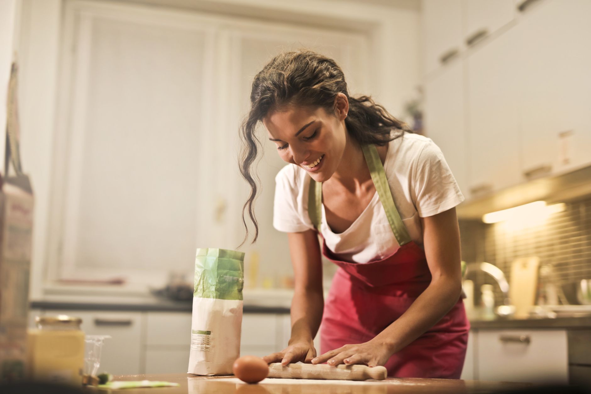 Low angle of cheerful young female in apron rolling dough at table while preparing food in modern light kitchen